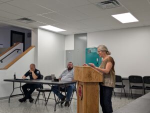 To the right, a woman stands behind a wooden podium. To the left, two men sit behind a folding table.